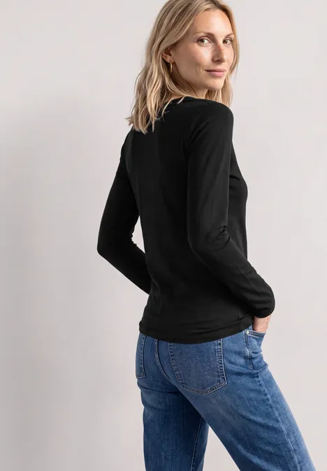 Frau in schwarzem Langarmshirt und blauen Jeans, seitlich stehend, den Kopf über die Schulter gewendet.