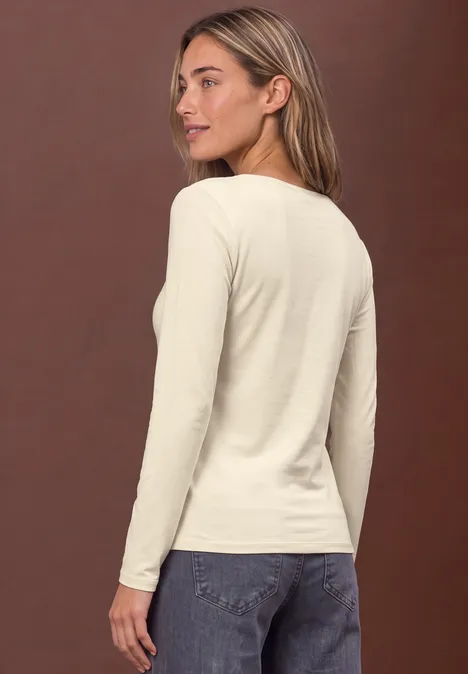 Vrouw gezien van achteren in crème lange mouw top en blauwe jeans voor een bruine achtergrond.