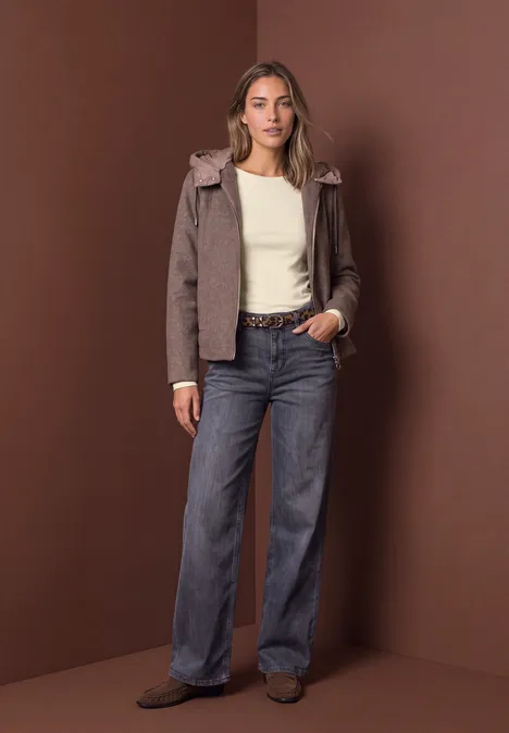 Vrouw met bruine jas, wit topje en wijde jeans, staand voor een bruin studio-achtergrond.