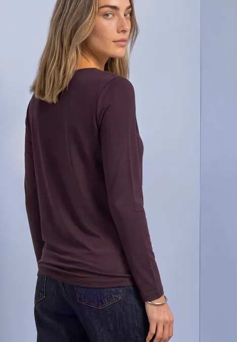 Jonge vrouw in bordeaux longsleeve en spijkerbroek, zijwaarts voor een effen achtergrond.