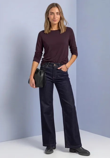 Vrouw in donkerrood trui, zwarte wijde jeans en schoenen, met zwarte schoudertas, voor grijs-paarse achtergrond.
