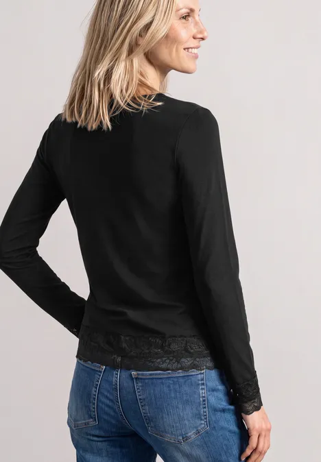 Frau von hinten in schwarzem Langarmshirt mit Spitzenbund und blauen Jeans, blondes Haar, lächelnd.