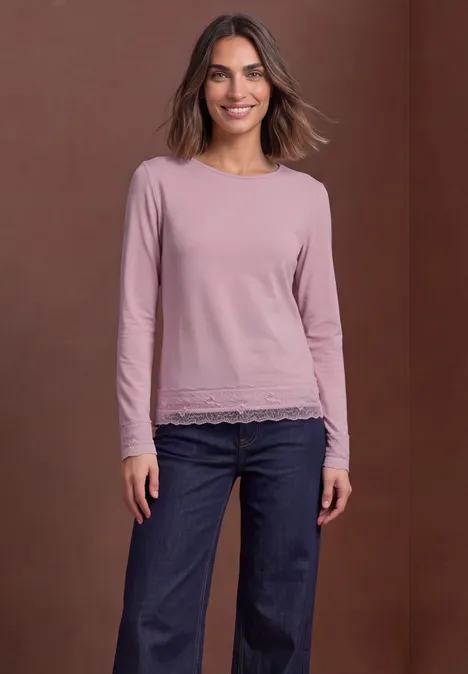 Vrouw lacht in een roze longsleeve met kanten rand en blauwe jeans voor een bruine achtergrond.