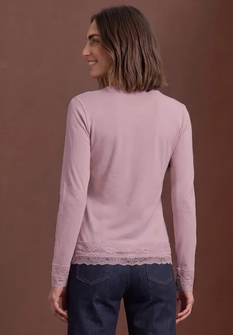 Een vrouw in een lichtroze top met kant en een spijkerbroek staat opzij voor een bruine achtergrond.