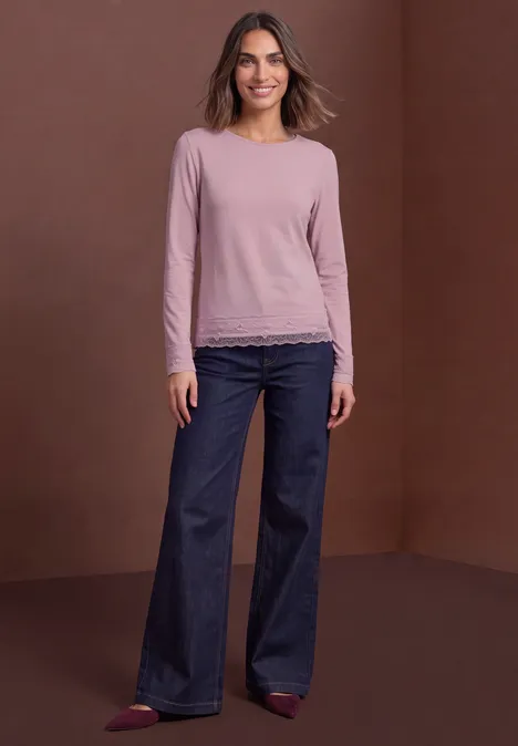 Vrouw in roze longsleeve met kant, wijde spijkerbroek en rode schoenen voor een bruine achtergrond.
