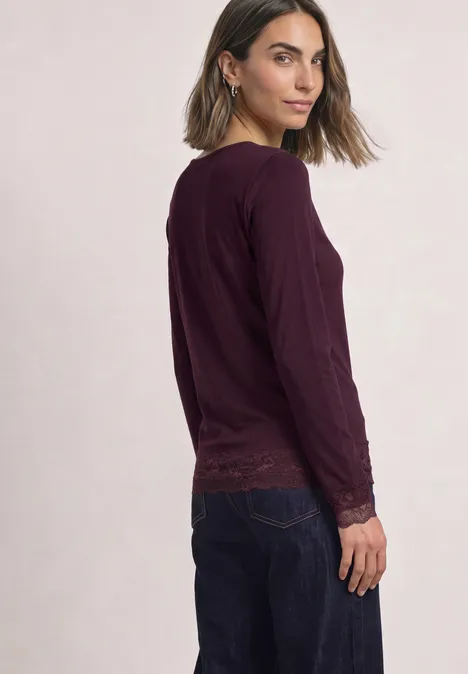 T-shirt à manches longues, col bateau et dentelle berry plum Frau im Profil, trägt dunkelviolette Langarmbluse mit Spitzendetails und dunkelblaue Jeans.