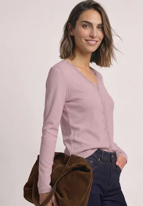 Veste à fines côtes, col en V et dentelle quiet rose Frau in rosa Langarmshirt, Jeans und brauner Tasche vor hellem Hintergrund.
