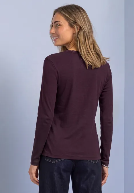 Eine Frau im dunkelvioletten Langarmshirt blickt lächelnd über die Schulter vor hellblauem Hintergrund.