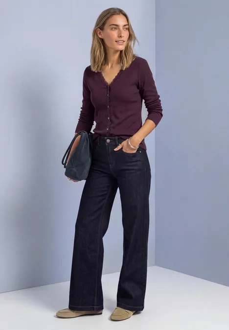 Eine Frau steht vor hellvioletter Wand, trägt eine bordeauxfarbene Strickjacke, weite Jeans und hält eine Clutch.