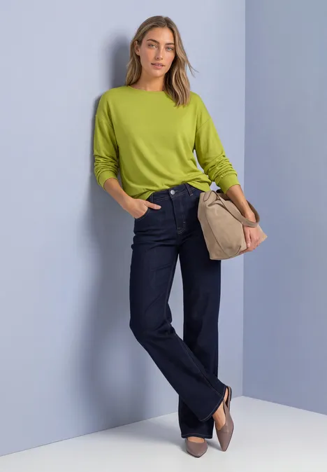 T-shirt en maille à manches chauve-souris apple lime Frau in leuchtend grünem Pullover, dunkelblauen Jeans und beiger Tasche steht vor hellem Hintergrund.