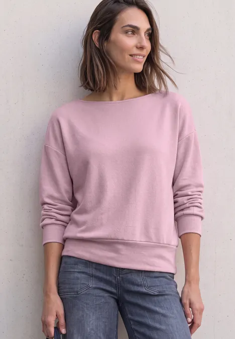 T-shirt en maille à manches chauve-souris quiet rose Frau steht lässig vor Wand, trägt rosa Sweatshirt und Jeans, Blick seitlich, entspannte Pose.