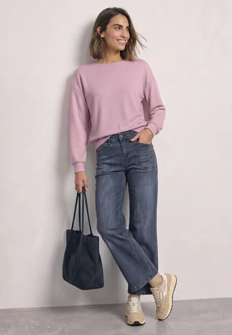 T-shirt en maille à manches chauve-souris quiet rose Frau in rosa Pullover, geraden Jeans und Turnschuhen trägt eine blaue Tasche und lächelt.