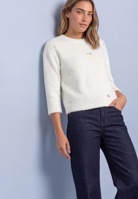3/4-Arm Shirt mit dekorativen Details off white Frau trägt weißen Lochstrickpullover mit 3/4-Ärmeln und dunkle Jeans vor pastellblauem Hintergrund.