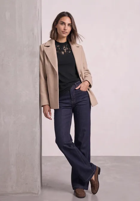 Top mit Rundhals und Spitze Black Eine Frau lehnt an einer Säule, trägt beige Blazer, schwarze Spitzenbluse und dunkle Jeans.