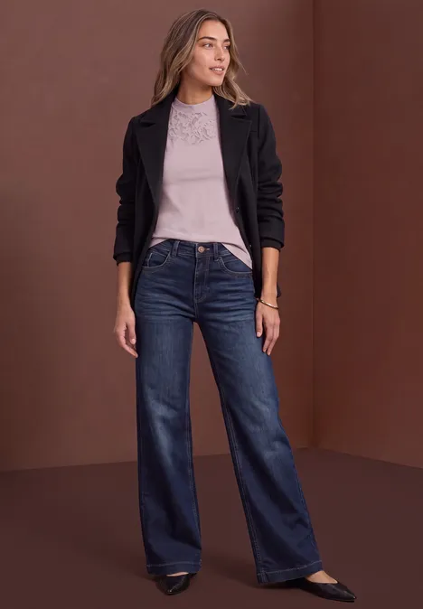 Top mit Rundhals und Spitze quiet rose Eine Frau in schwarzer Jacke, grauem Shirt und weiten Jeans posiert vor braunem Hintergrund.