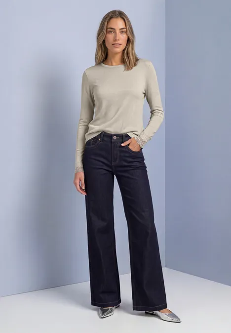 T-shirt à manches longues avec paillettes et dentelle pure beige Frau in sportlich-elegantem Outfit: helles Langarmshirt, weite dunkle Jeans und spitze Schuhe vor neutralem Hintergrund.