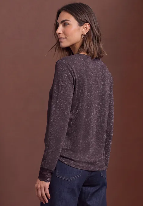 Frau in violettem, langärmligem Pullover und Jeans, seitlich abgewandt vor braunem Hintergrund.