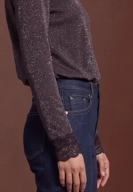 Seitenansicht einer Person im glitzernden Langarmshirt mit Spitzenärmeln und blauen Jeans, brauner Hintergrund.
