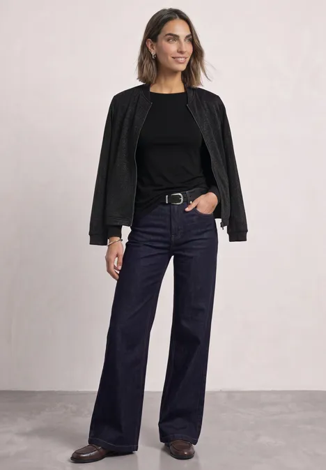 Frau in schwarzer Jacke, schwarzem Shirt und weitem, dunklen Jeanshose vor hellem Hintergrund.