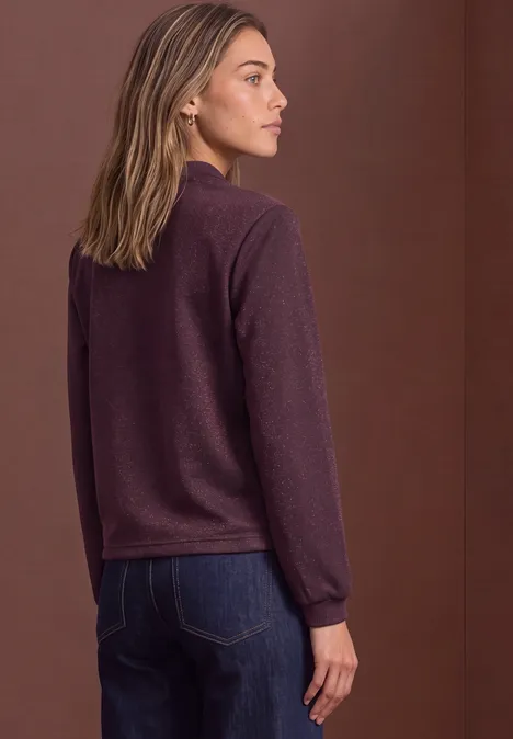 Seitlich stehende Frau in bordeauxrotem Pullover und Jeans vor braunem Hintergrund.