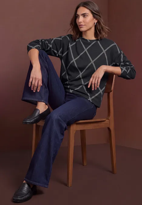 T-shirt à manches longues, col rond et motif à carreaux armor grey Femme assise sur une chaise, porte un pull gris foncé à motifs quadrillés blancs, jean bleu foncé et mocassins noirs.