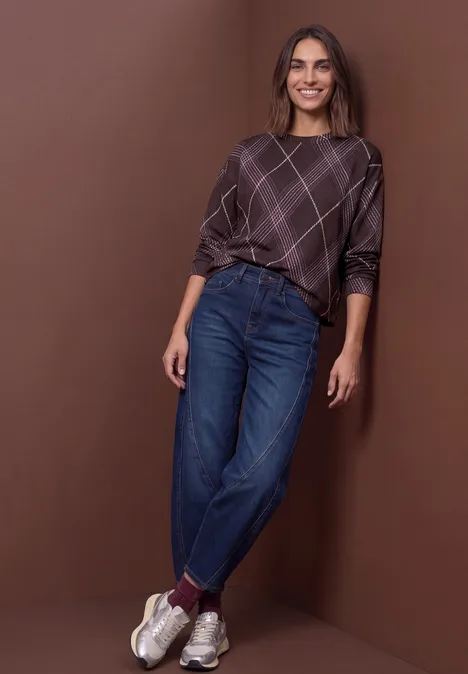 T-shirt à manches longues, col rond et motif à carreaux berry plum Femme avec un pull à carreaux, jean bleu et baskets blanches devant un fond marron, souriante et décontractée.