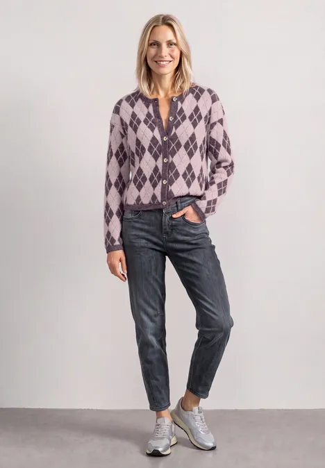 Frau in argylegemustertem Pullover, Jeans und Sneakern vor neutralem Hintergrund.