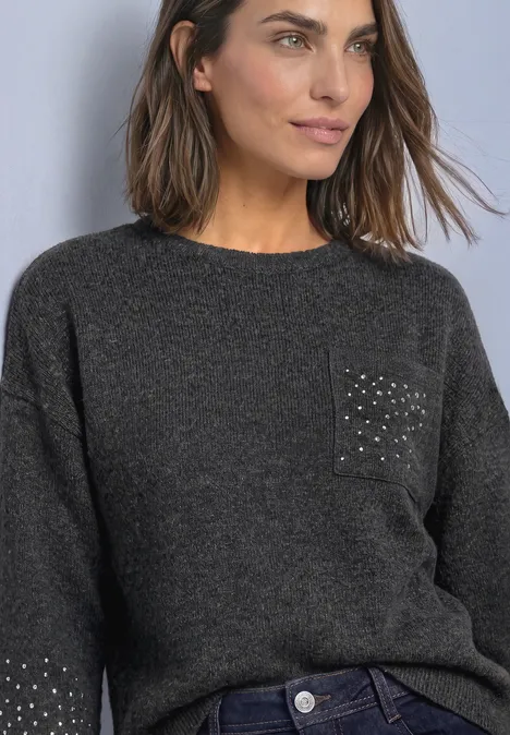 Frau in grauem Strickpullover mit Nieten auf der Tasche und Jeans, schaut seitlich.