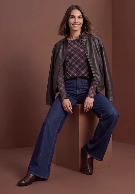 Vrouw met leren jas over schouders, ruitjestrui, hoge flare-jeans en bruine enkellaarsjes.
