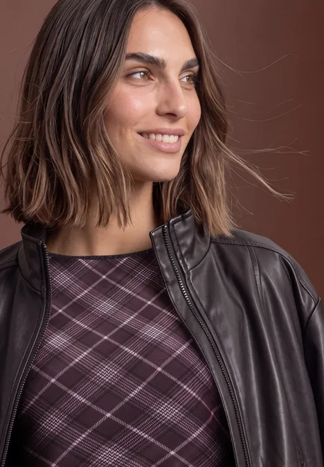 Een vrouw glimlacht zijwaarts, gekleed in een bruine leren jas met paars ruitjesshirt.