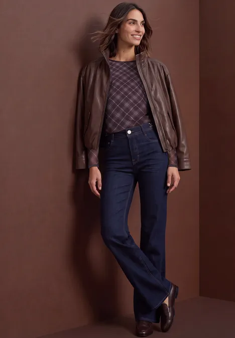 Vrouw met bruine leren jas, gebreide trui, donker flared jeans en laarzen voor een bruine achtergrond.