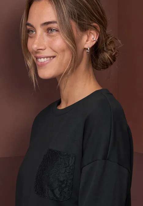 Junge Frau mit zurückgebundenem Haar, lächelnd, trägt schwarzen Pullover mit Spitzentasche.