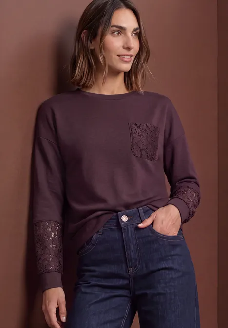 Frau in lila Langarmshirt mit Spitzenbesatz an Tasche und Ärmeln, dunkle Jeans, vor braunem Hintergrund.