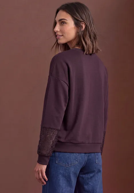 Frau in violettem Pullover mit Spitzenärmeln und blauer Jeans, Blick über die Schulter, brauner Hintergrund.