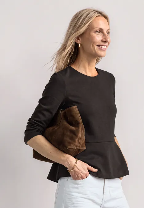 Frau in schwarzem Peplum-Top und weißen Hosen, lächelt und hält eine braune Clutch vor hellem Hintergrund.