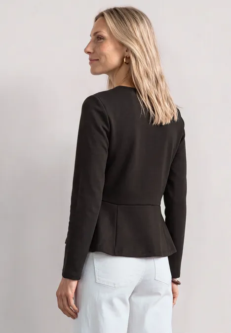 Eine blonde Frau von hinten, trägt schwarzen Blazer und weiße Jeans, steht seitlich und blickt zur Seite.