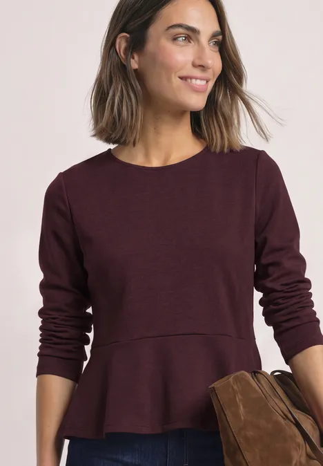 Frau mit brauner Tasche trägt burgunderfarbenes Langarmshirt mit Schößchen-Detail und lächelt.
