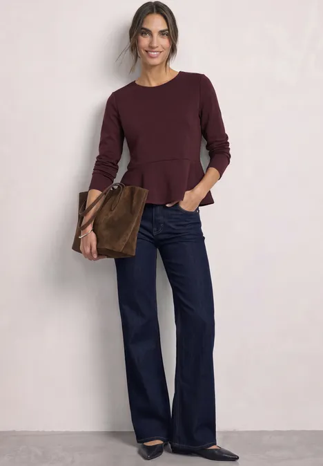 Frau in burgunderrotem Peplum-Oberteil, weiten Jeans und brauner Tasche vor neutralem Hintergrund.