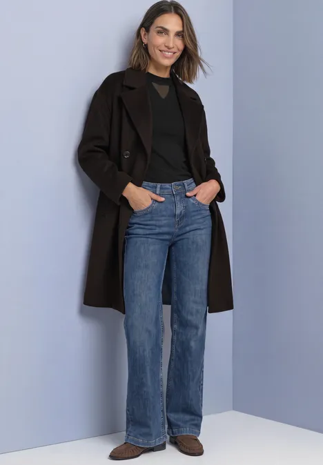 Frau mit braunem Mantel, schwarzem Shirt, blauen Jeans und braunen Schuhen vor hellem Hintergrund.