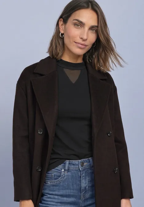 Junge Frau in schwarzem Top, dunklem Mantel und Jeans vor neutralem Hintergrund.