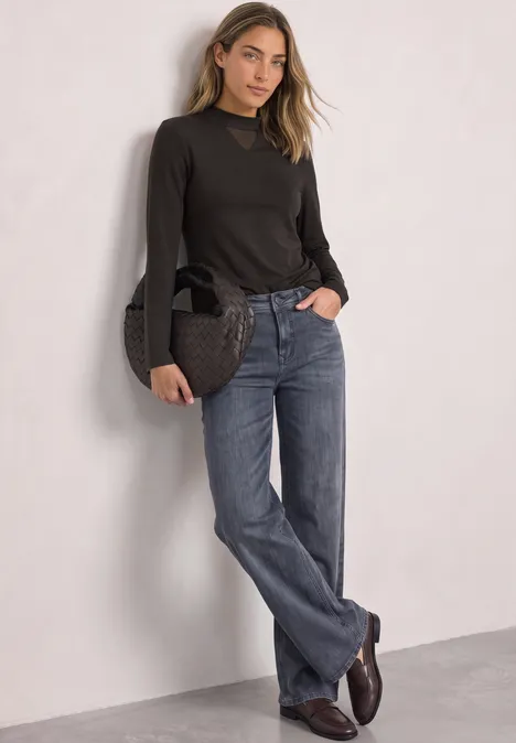 Frau in dunkelbraunem Oberteil und blauen Jeans lehnt an heller Wand und hält eine Handtasche.