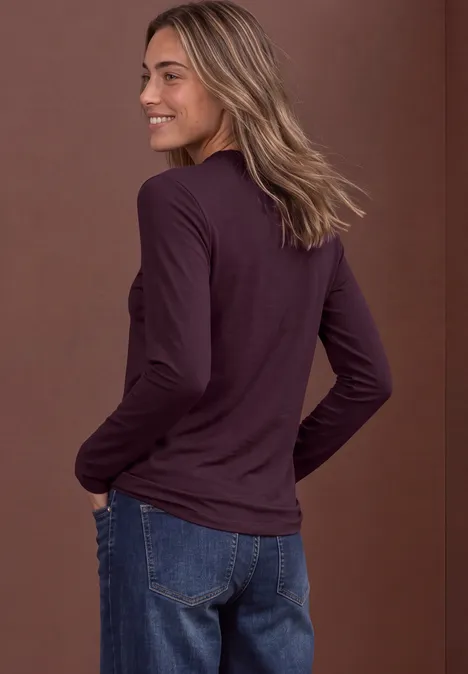 Eine lächelnde Frau im Profil trägt ein langärmeliges, dunkelviolettes Shirt und blaue Jeans.