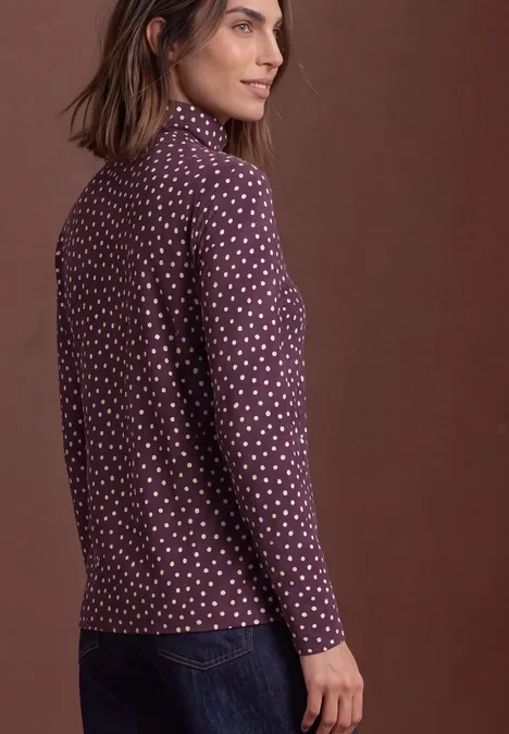 Seitliche Ansicht einer Frau in lila, gepunktetem Langarmshirt und Jeans vor braunem Hintergrund.