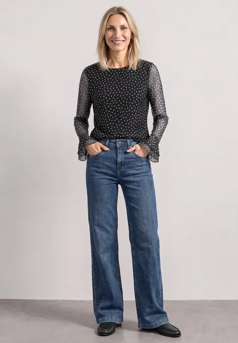 Frau in schwarz-weiß gepunkteter Bluse mit Rüschenärmeln und weiten blauen Jeans schwarze Schuhe vor grauem Hintergrund.