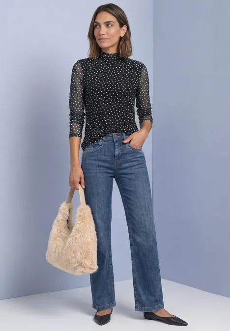 Frau mit schwarzer, gepunkteter Bluse, blauen Jeans und flauschiger Tasche vor schlichtem Hintergrund.