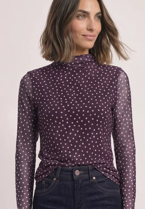 Frau in violettem, gepunktetem Langarm-Transparenzshirt mit Stehkragen, dazu dunkle Jeans.