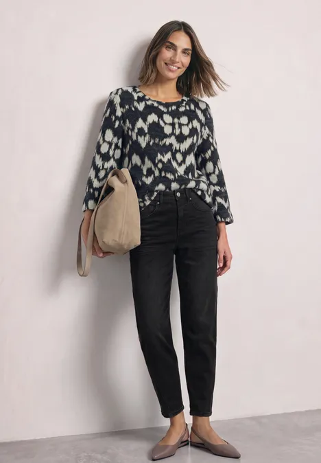 Een lachende vrouw draagt een zwarte high-waist jeans, een gemusterd topje en heeft een beige tas.