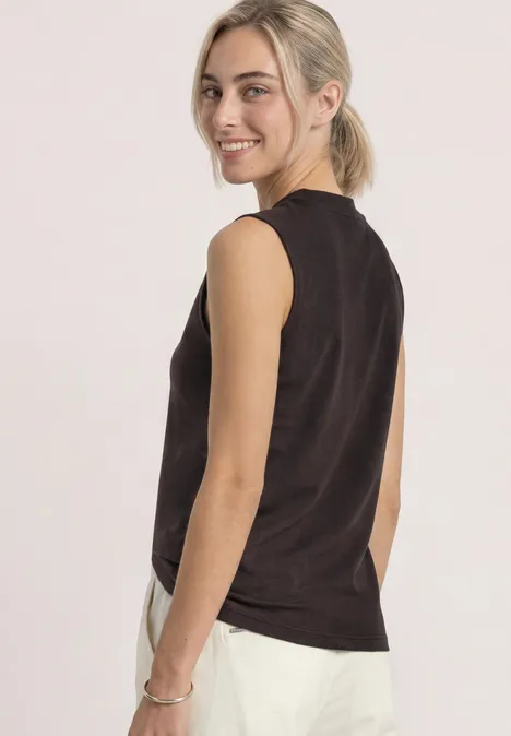 Jeune femme blonde en queue de cheval, débardeur noir et pantalon blanc, souriant par-dessus son épaule.