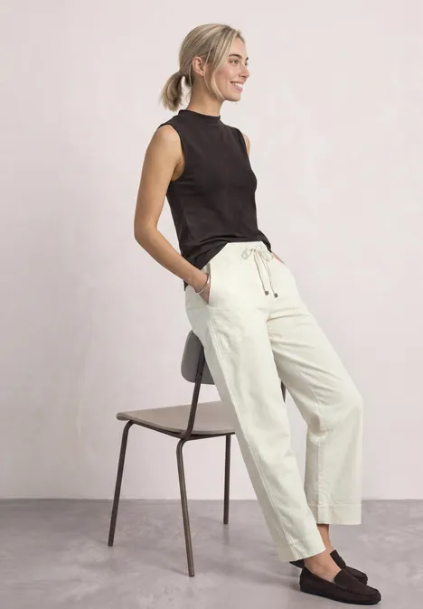Femme en top noir et pantalon blanc, adossée à une chaise, posture décontractée.