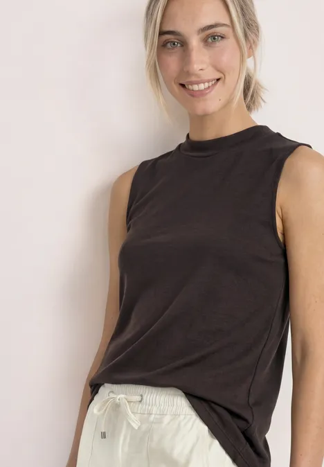 Une femme souriante porte un haut sans manches noir et un pantalon blanc sur fond neutre.